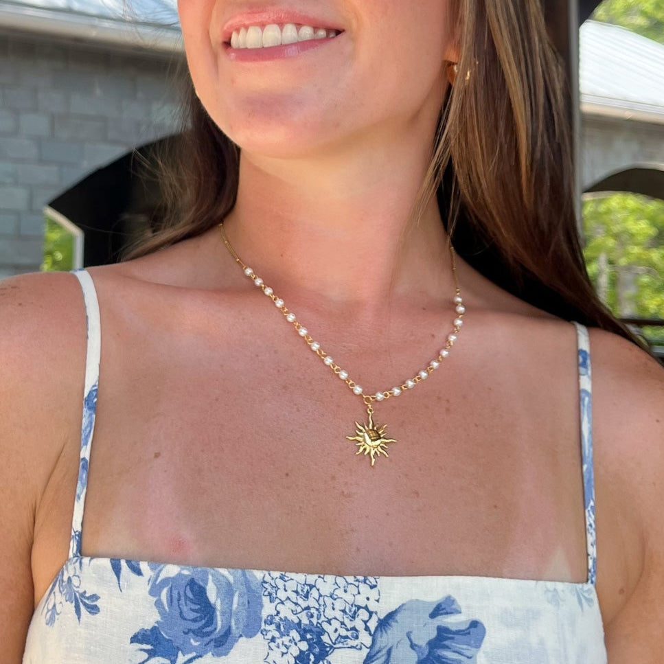 Woman wearing a floral dress and gold necklace with a sun design outdoors.